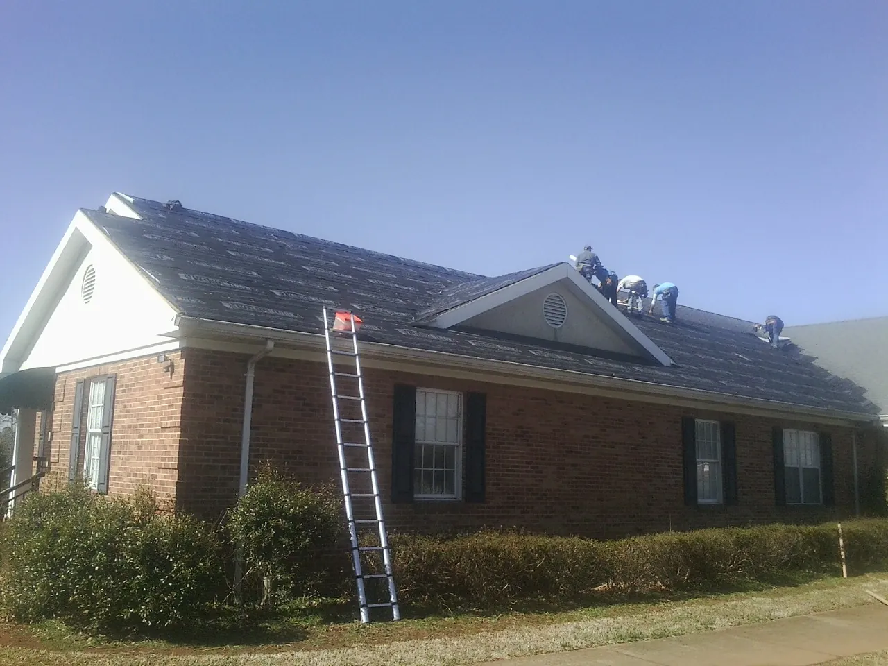 Crew tearing shingles off a brick ranch roof with a ladder set — Metal Roof Repair work by our crew in Dana